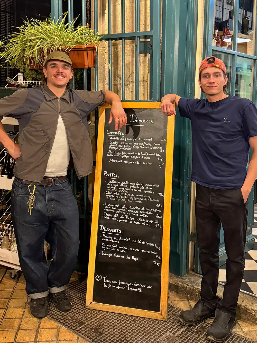 In einem Restaurant mit gefliestem Boden und Glasfenstern stehen zwei Personen lächelnd neben einer großen Kreidekarte.