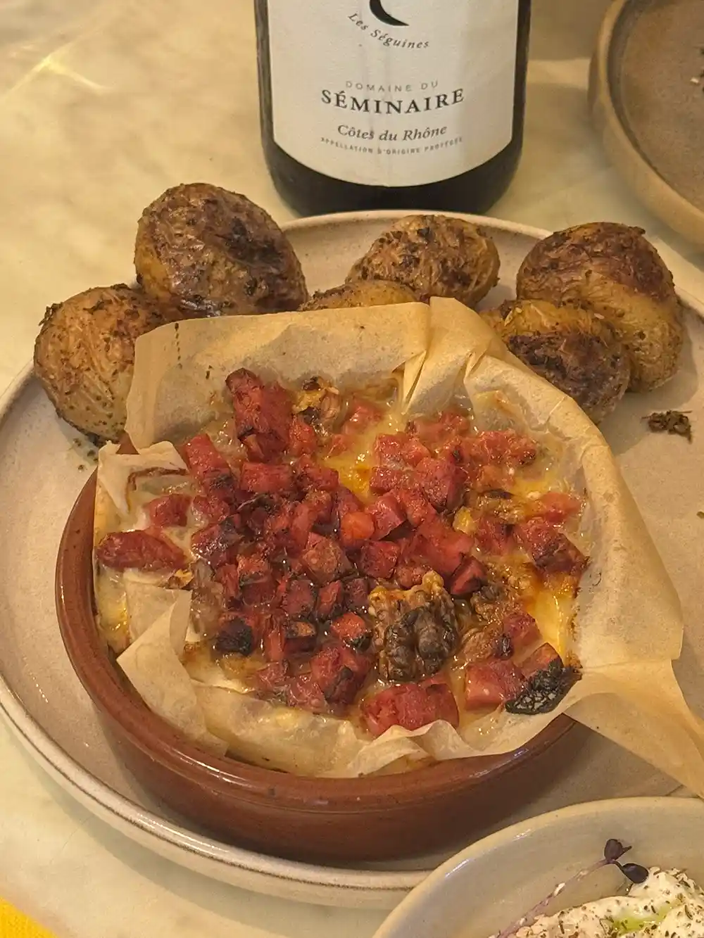 Überbackener Käse mit Wurstwürfeln, serviert mit Bratkartoffeln und einer Flasche Wein im Hintergrund.