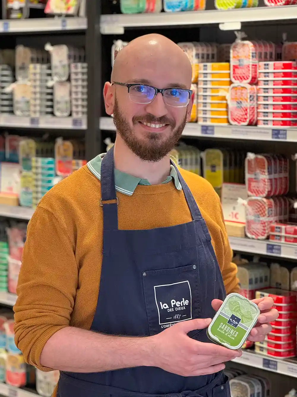Ein lächelnder Mann in einer Schürze hält eine Dose Fisch in einem mit Konserven gesäumten Gang eines Geschäfts.