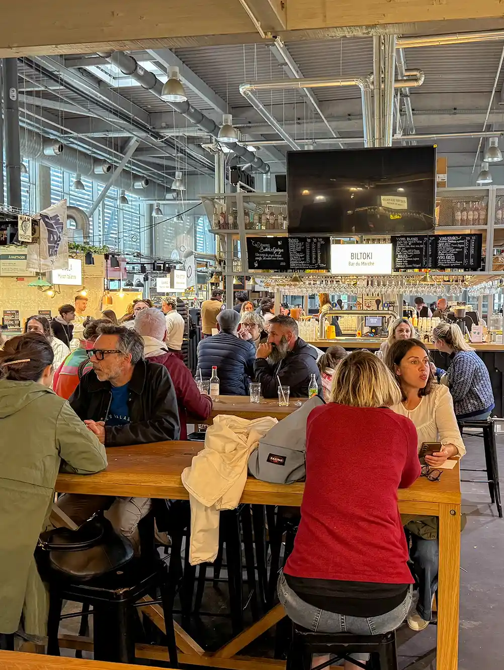 Menschen sitzen an Holztischen, essen und unterhalten sich in einer belebten, industriell anmutenden Lebensmittelhalle.