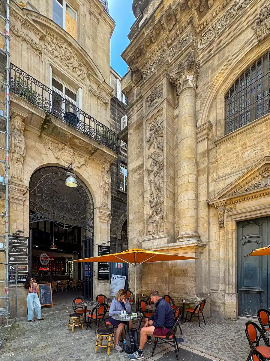 Bordeaux-Geheimtipps-Notre-Dame-Teeladen An einem sonnigen Tag sitzen die Menschen an Cafétischen im Freien zwischen kunstvoller Steinarchitektur und großen Torbögen.