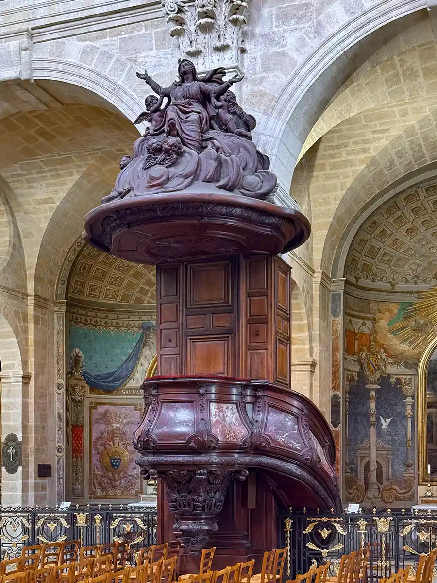Bordeaux-Geheimtipps-Notre-Dame-de-Bordeaux-Kanzel Verschnörkelte Holzkanzel mit detaillierten Schnitzereien vor Steinbögen und farbenfroher Wandkunst.