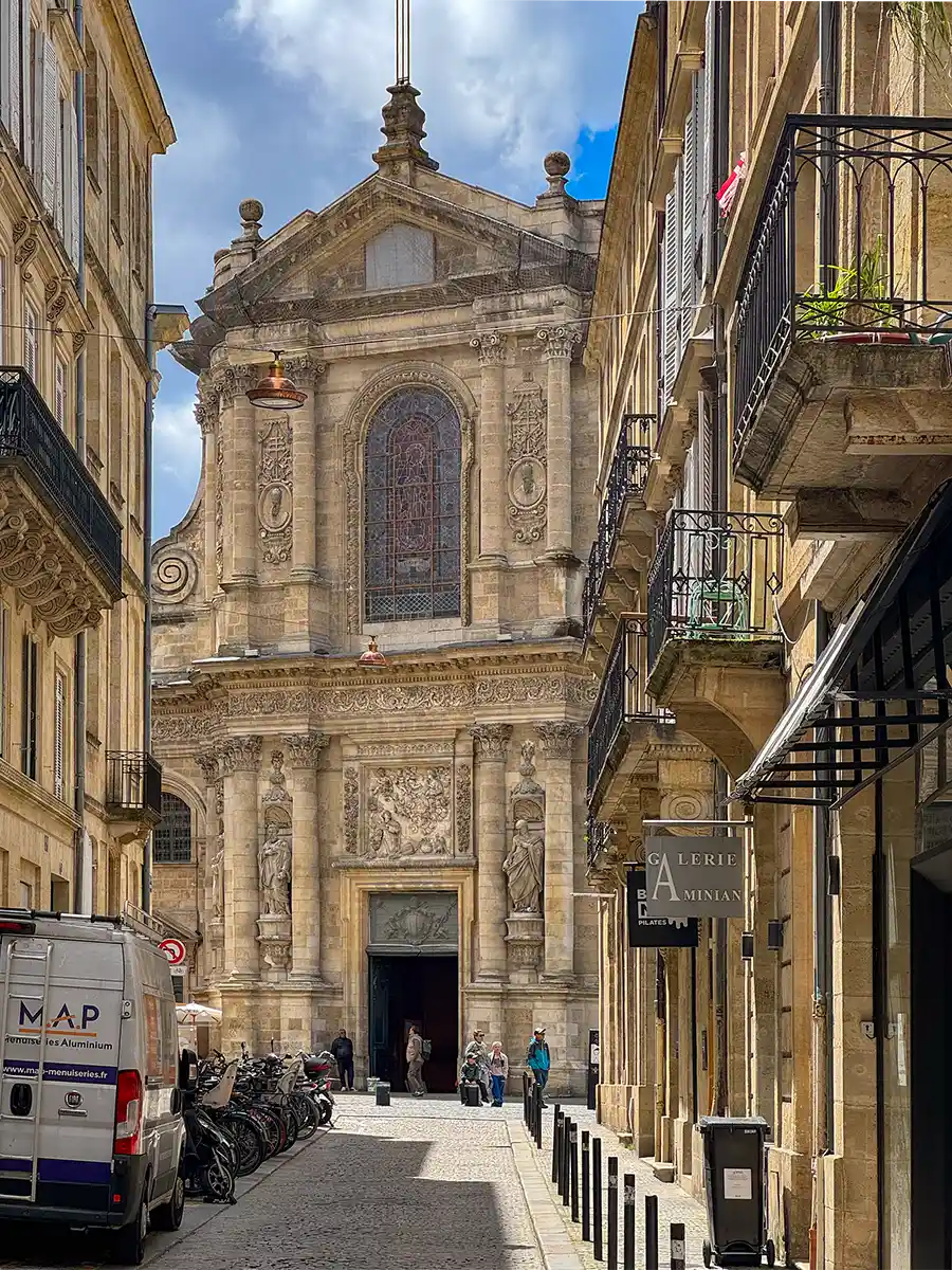Bordeaux-Geheimtipps-Notre-Dame-de-Bordeaux Eine historische Steinkirche steht am Ende einer schmalen Straße, die von Balkonen und abgestellten Fahrrädern gesäumt ist.