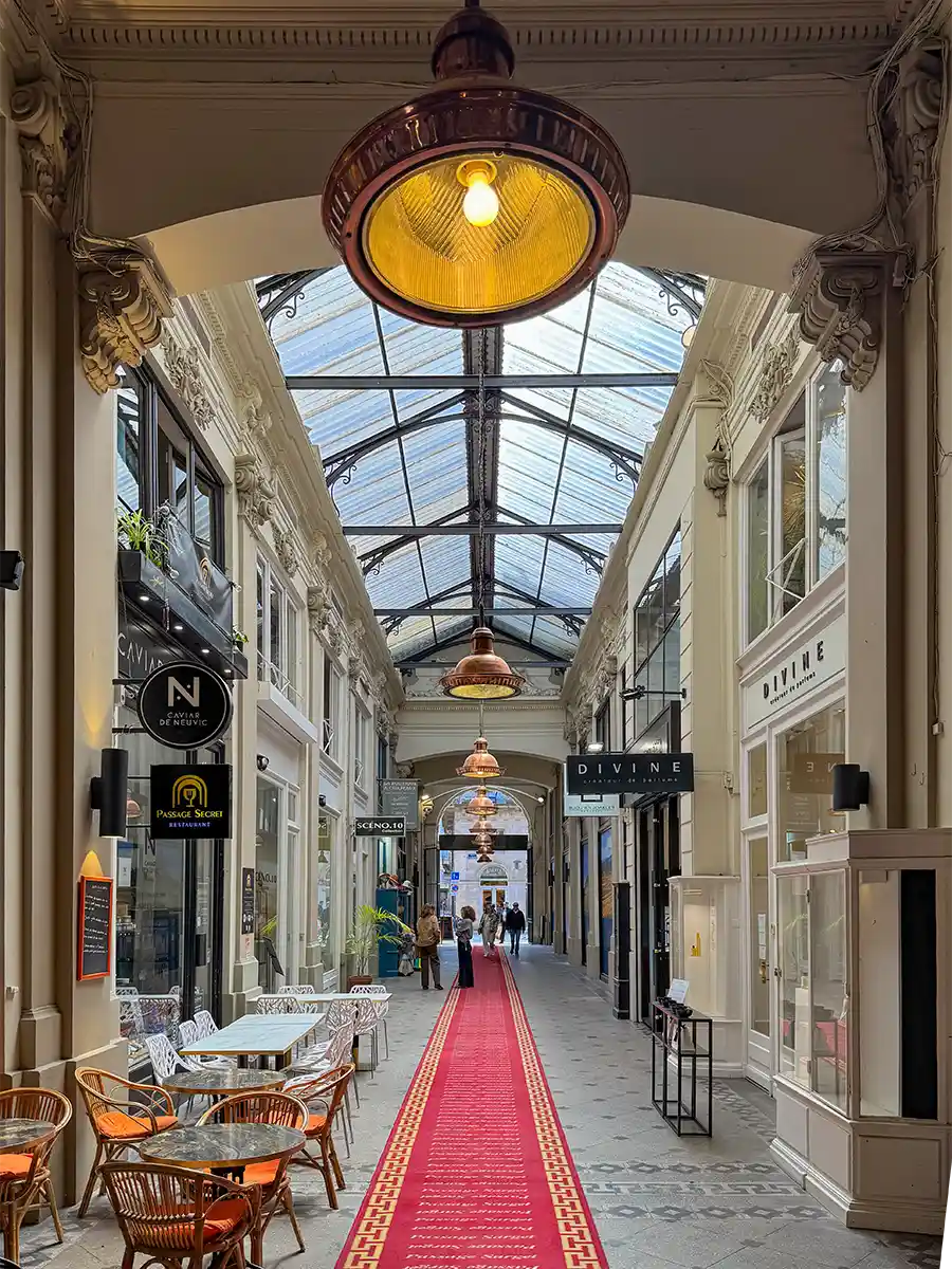 Bordeaux-Geheimtipps-Passage-Sarget Eine glasüberdachte Einkaufspassage mit einem roten Teppich, Geschäften und einem kleinen Café mit Terrassenstühlen auf der linken Seite.