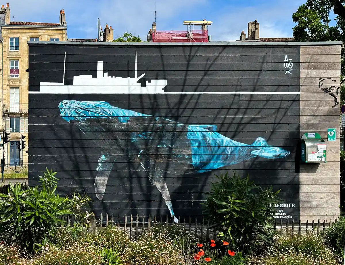 Bordeaux-Geheimtipps-Street-Art-Afrique Wandgemälde eines Blauwals unter Wasser mit einem weißen Schiff darüber; vor dem Wandgemälde wachsen Pflanzen.