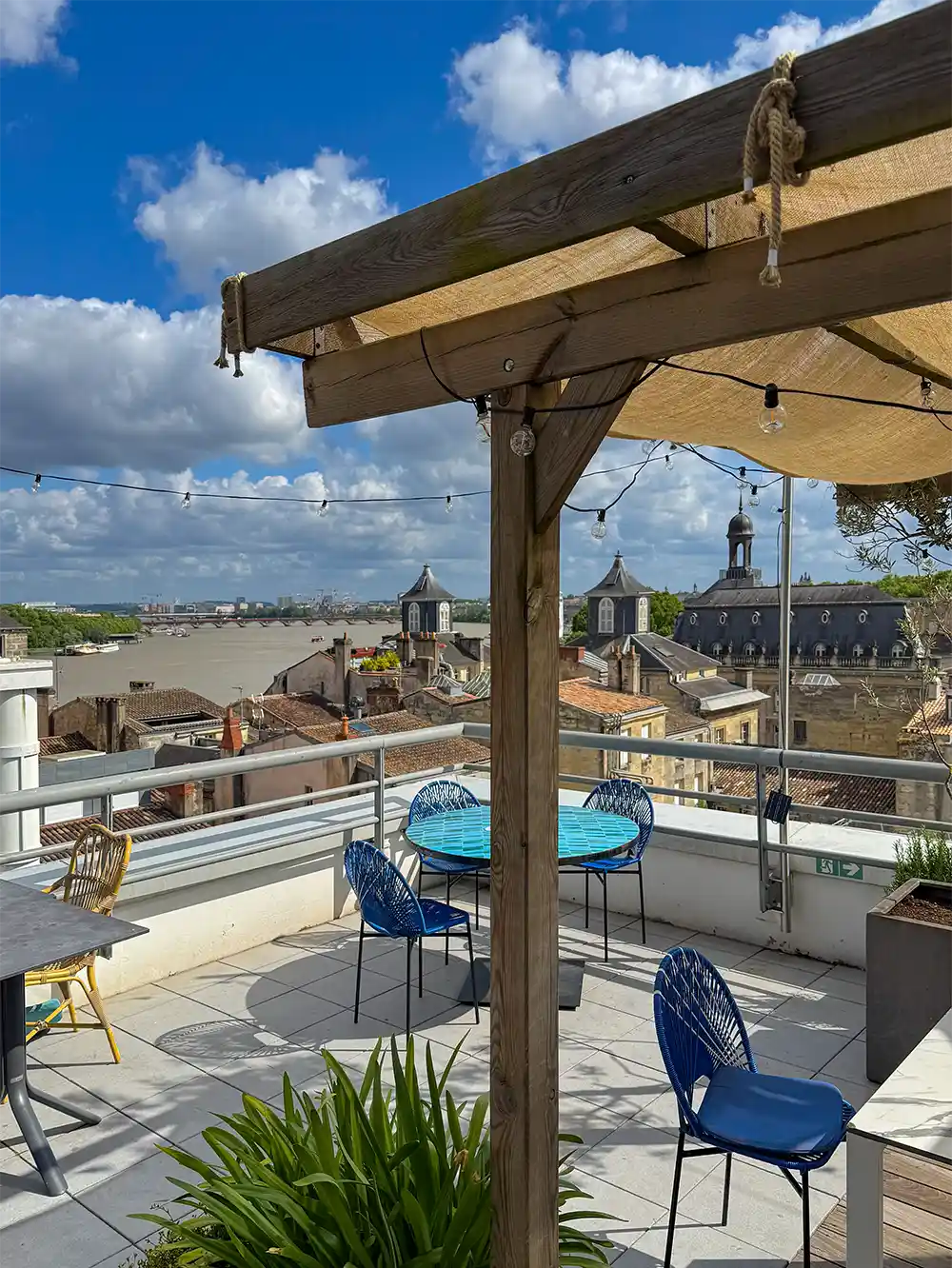 Dachterrasse mit blauen Stühlen, Tischen, Lichterketten und Blick auf die Stadt unter einem teilweise bewölkten Himmel.