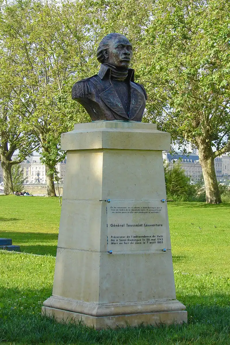 Bordeaux-Geheimtipps-Toussaint-Louverture Bronzebüste eines Mannes auf einem Steinsockel in einem grasbewachsenen Park mit Bäumen und Gebäuden im Hintergrund.