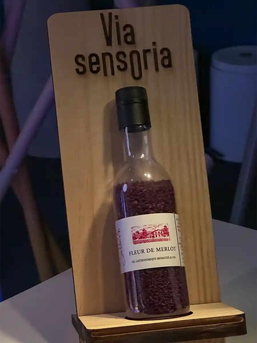 Bordeaux-Geheimtipps-Via-Sensoria-Fleur-de-Merlot Eine Flasche mit der Aufschrift Fleur de Merlot mit violetter Maserung, die auf einem Holzständer mit der Aufschrift Via sensoria steht.
