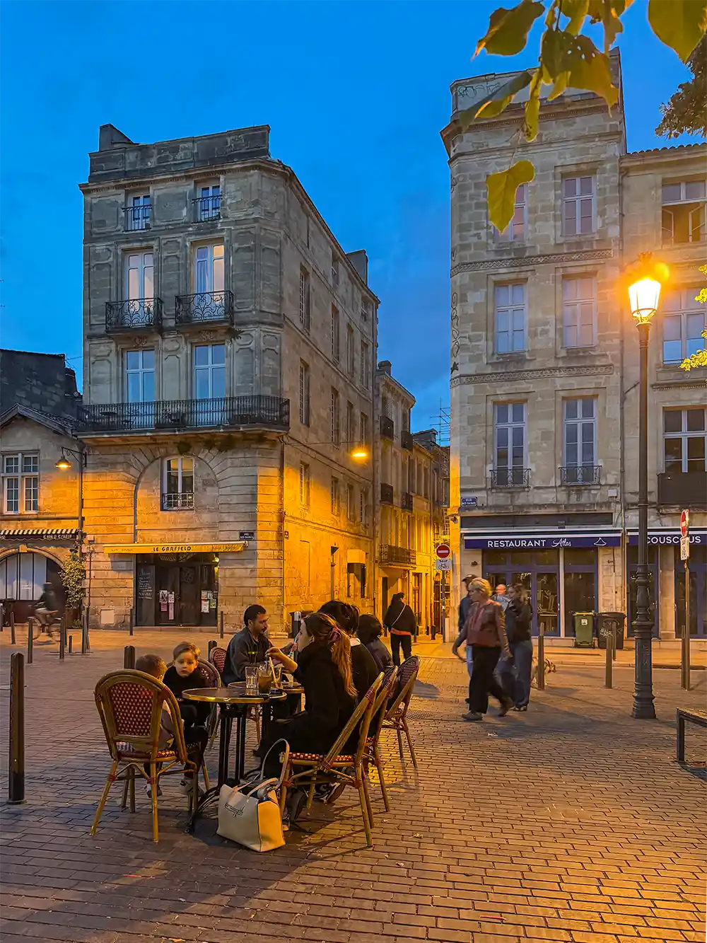 Menschen, die in einem Straßencafé auf einem europäischen Stadtplatz in der Abenddämmerung mit historischen Gebäuden und Straßenlaternen speisen.