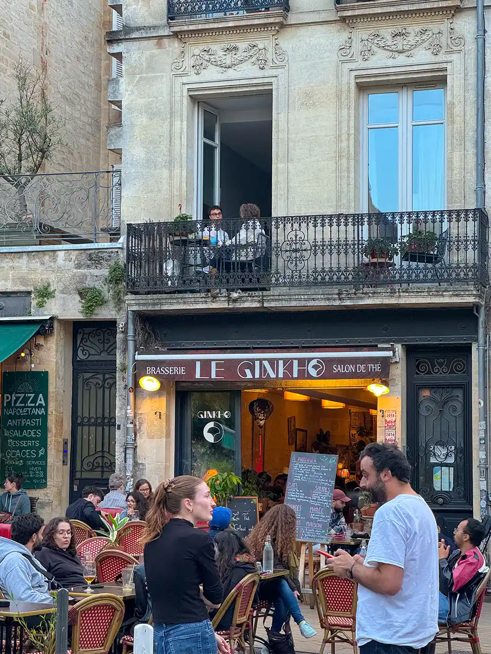 Menschen essen in einem Café im Freien, einige sitzen auf einem Balkon vor einem Gebäude.