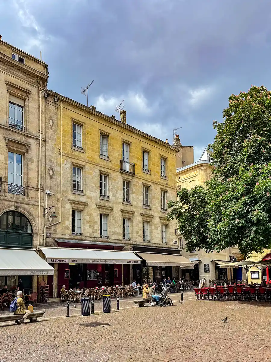 Bordeaux-Place-St-Pierre Ein kopfsteingepflasterter Platz mit Straßencafés, sitzenden Menschen und historischen Steingebäuden unter einem bewölkten Himmel.