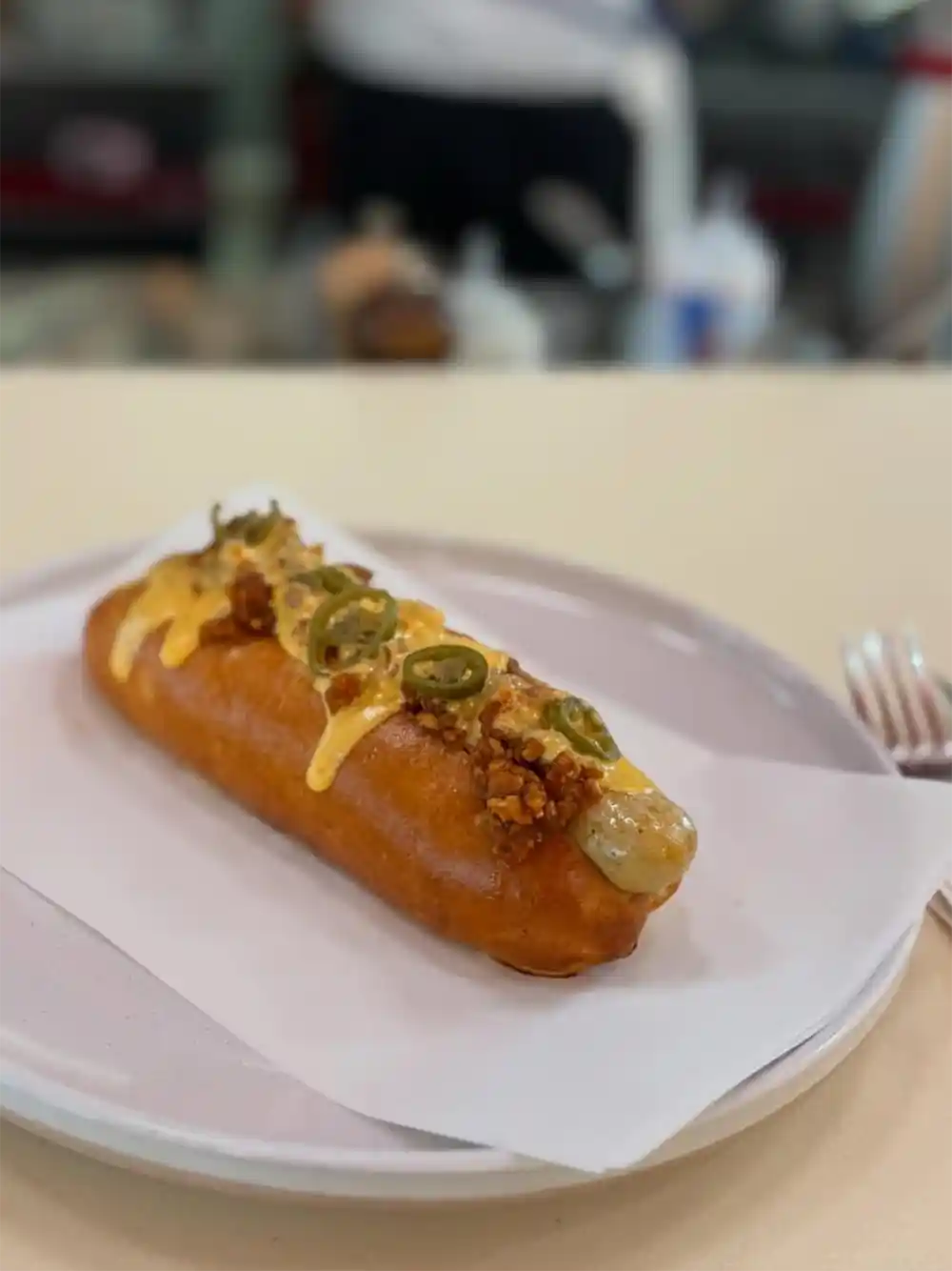 Ein frittierter Hot Dog, der mit geschmolzenem Käse, Jalapeños und Streuseln belegt und auf einem weißen Teller serviert wird.