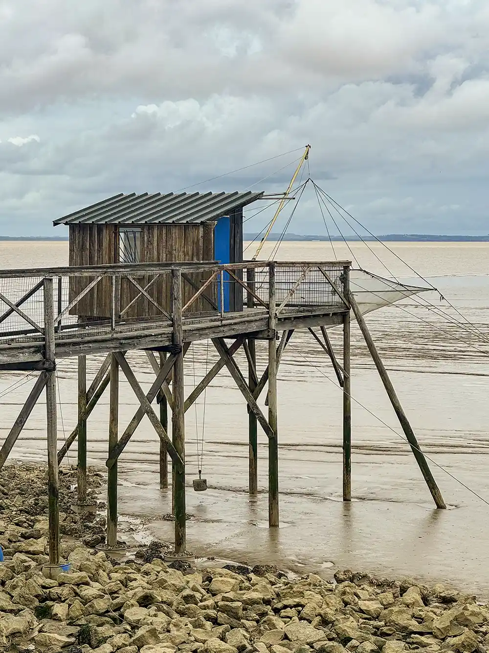 Carrelet-Gironde Eine hölzerne Fischerhütte auf Stelzen steht über einer felsigen Küste, über der sich Netze und ein bewölkter Himmel spannen.