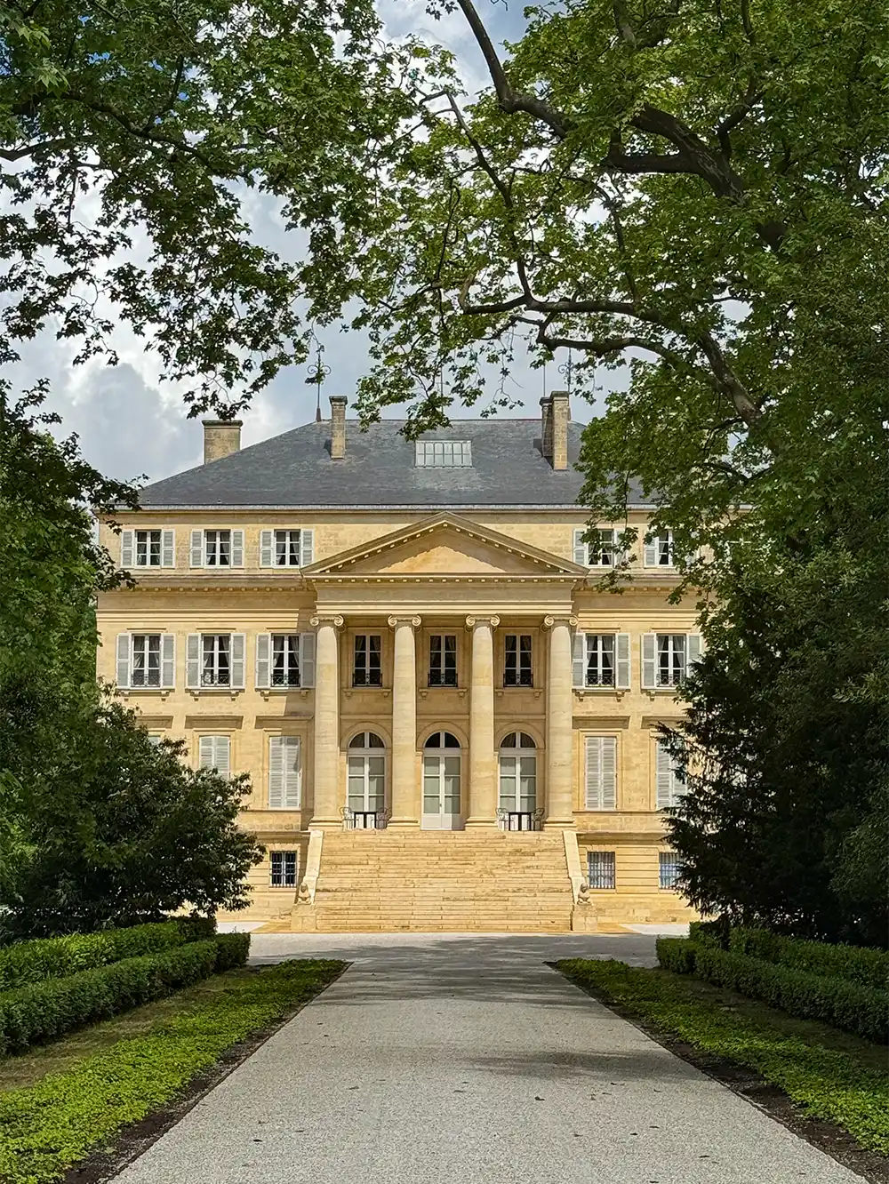 Ein großes steinernes Herrenhaus mit hohen Säulen, umgeben von Bäumen und Grün, das von einem Kiesweg aus zu sehen ist.