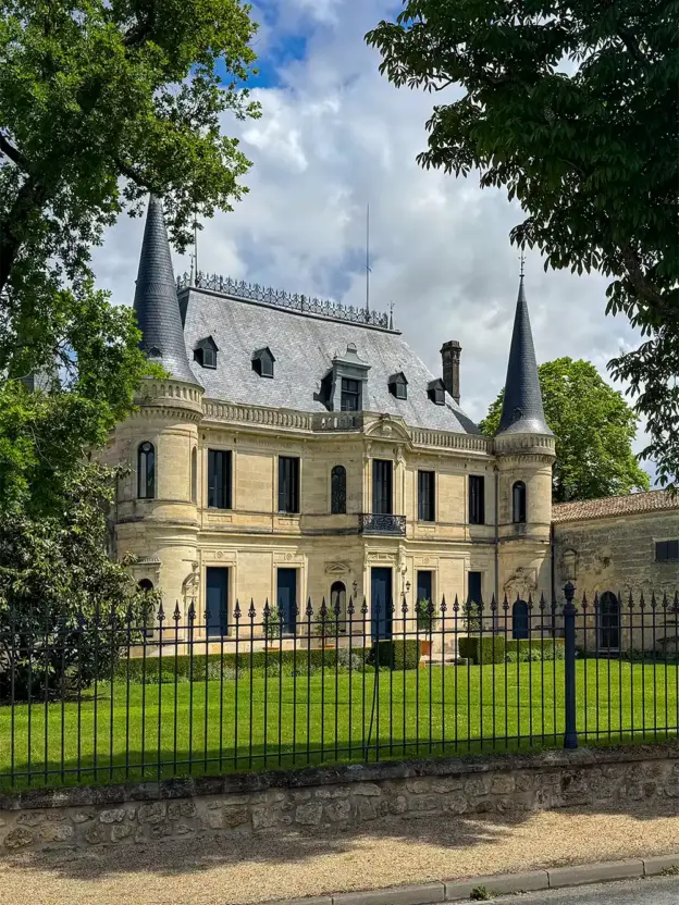 Ein steinernes Herrenhaus mit zwei spitzen Türmen, verziertem Dach und eingezäuntem Garten, umgeben von Bäumen.