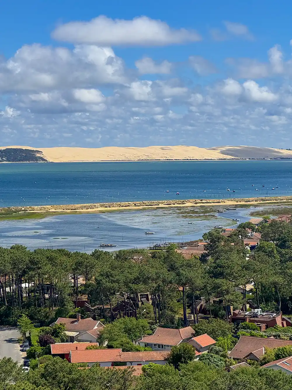Dune-de-Pilat-Arcachon Ein Küstendorf mit roten Dächern, grünen Bäumen, blauem Wasser und sandigen Dünen unter einem teilweise bewölkten Himmel.