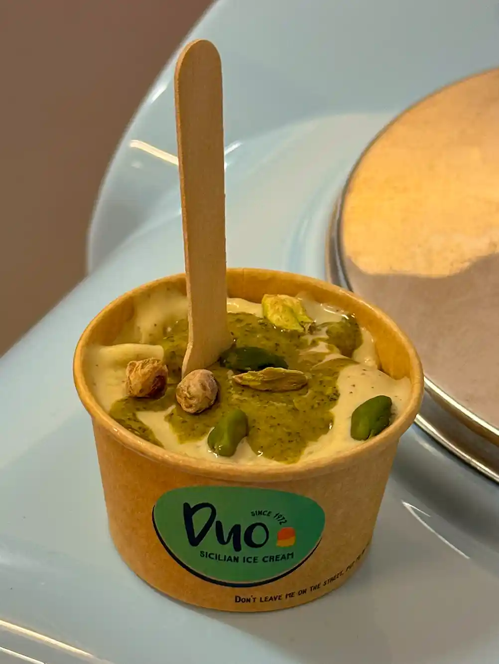 Duo-Gelato-Pistazien Ein Becher Pistazieneis mit ganzen Pistazien und einem Holzlöffel oben drauf.