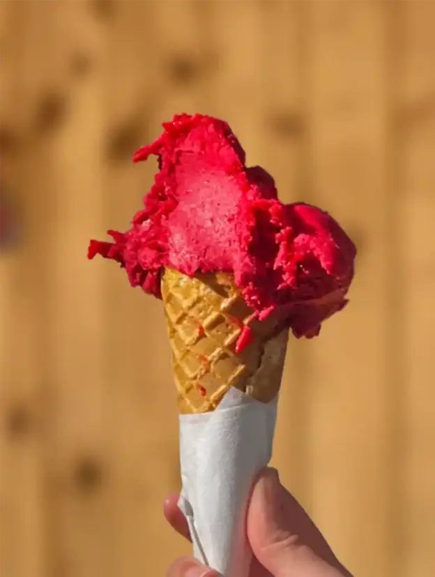 Eine Hand hält eine Waffeltüte mit leuchtend rosa Eis vor einem unscharfen braunen Hintergrund.
