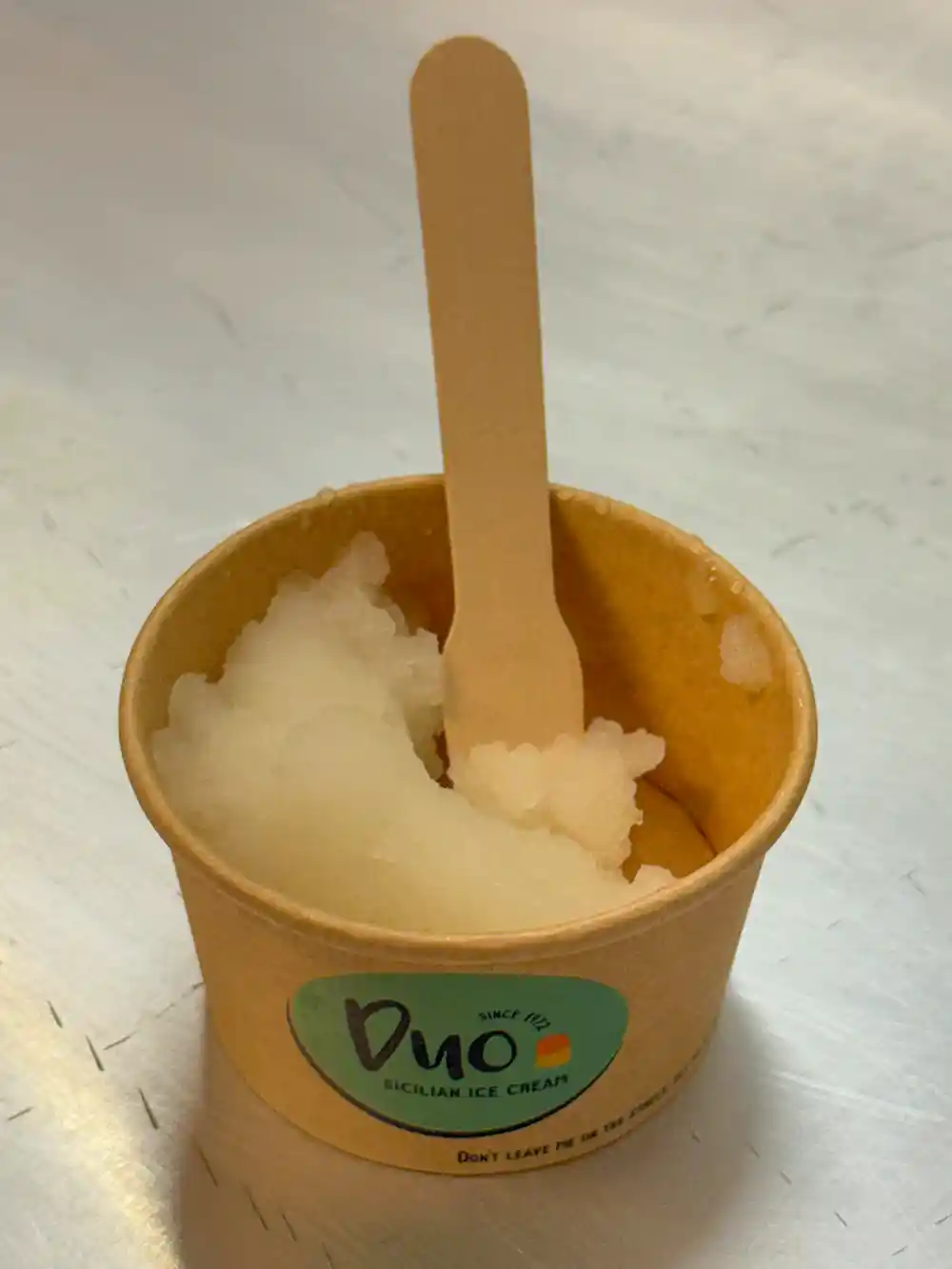 Duo-Zitronen-Granita Ein kleiner Pappbecher mit weißem sizilianischem Eis und einem Holzlöffel auf einer Metalloberfläche.