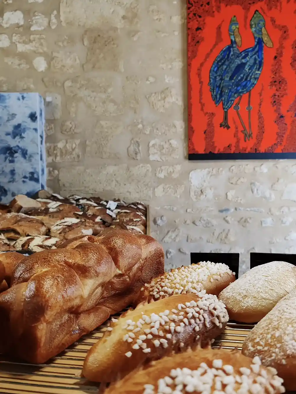 Geheimtipps-Bordeaux-Ceres-Boulangerie Verschiedene Brotsorten auf einem Gestell mit einer Backsteinmauer und einem bunten Vogelgemälde im Hintergrund.