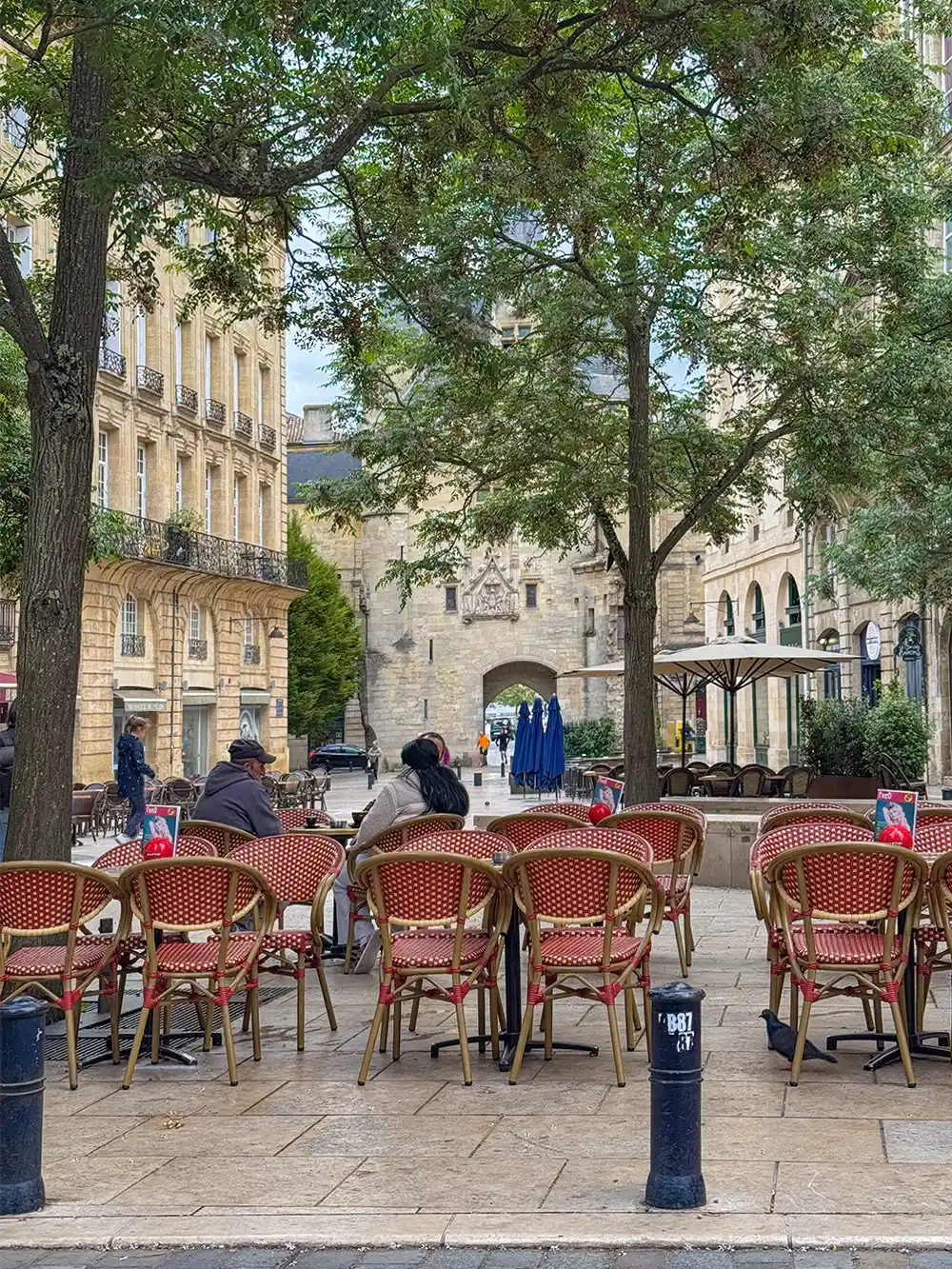 Geheimtipps-Bordeaux-Chez-Fred-Porte-Cailhau Außencafé mit roten Stühlen, Menschen sitzen unter Bäumen, historisches Steingebäude im Hintergrund.