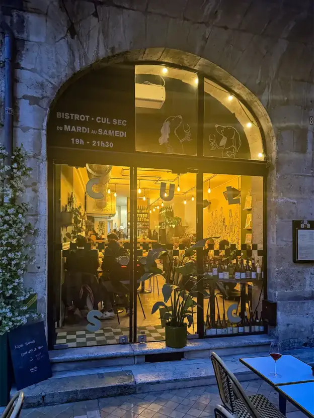 Ein gemütliches, warm beleuchtetes Bistro, in dem nachts durch große Bogenfenster gegessen wird.