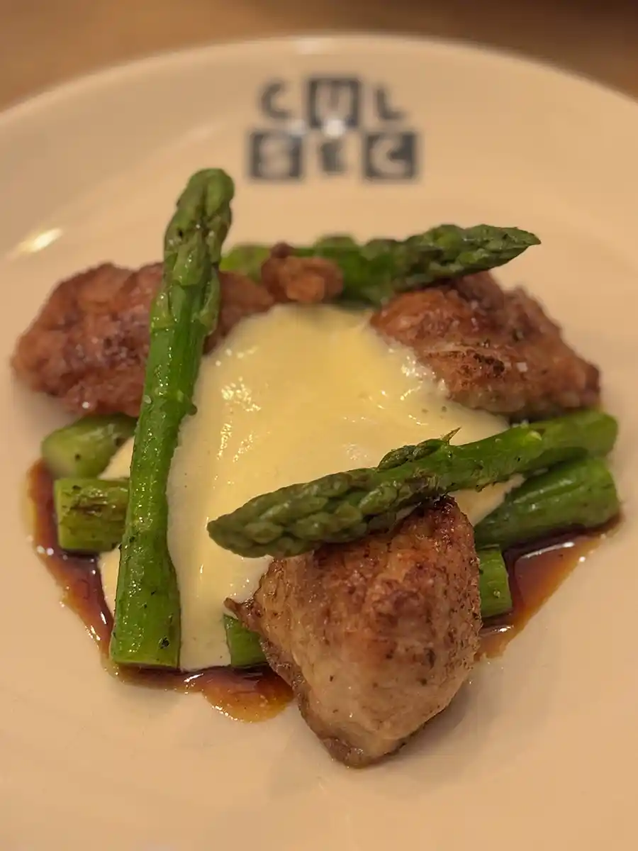 Geheimtipps-Bordeaux-CulSec-Kalbsbries-Spargel Hähnchenteile mit Spargelstangen und Sahnesauce, serviert auf einem weißen Teller mit einem Logo am Rand.