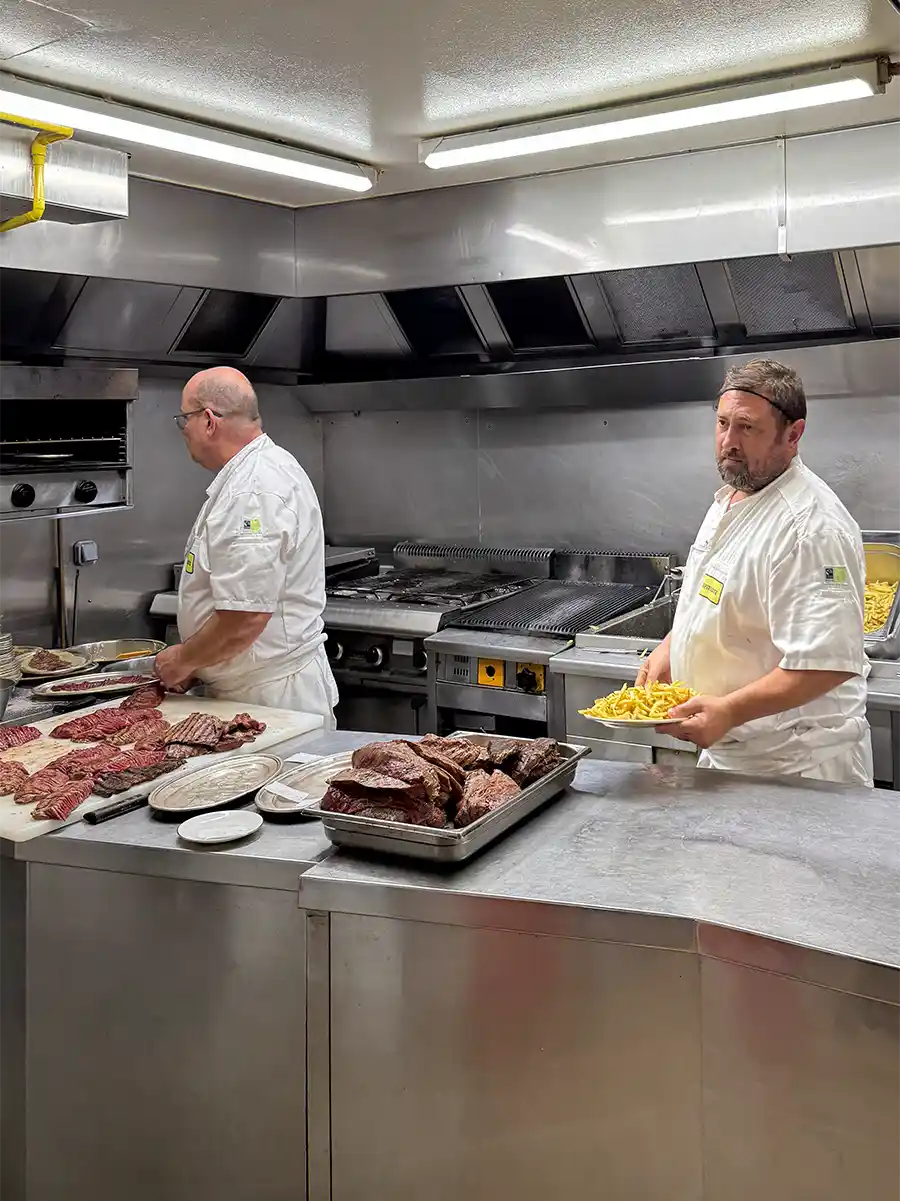 Geheimtipps-Bordeaux-Restaurant-L-Entrecote-Kueche Zwei Köche bereiten in einer Großküche Fleisch und Pommes frites auf Arbeitsflächen und Tabletts aus Edelstahl zu.
