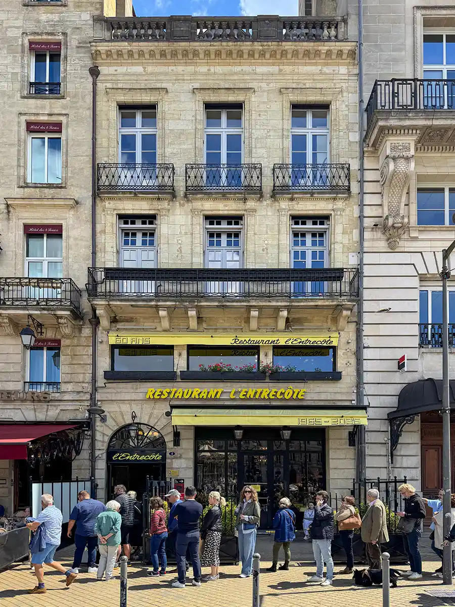 Geheimtipps-Bordeaux-Restaurant-L-Entrecote Menschen stehen vor dem Restaurant L'Entrecôte, das in einem klassischen mehrstöckigen Steingebäude mit Balkonen untergebracht ist.