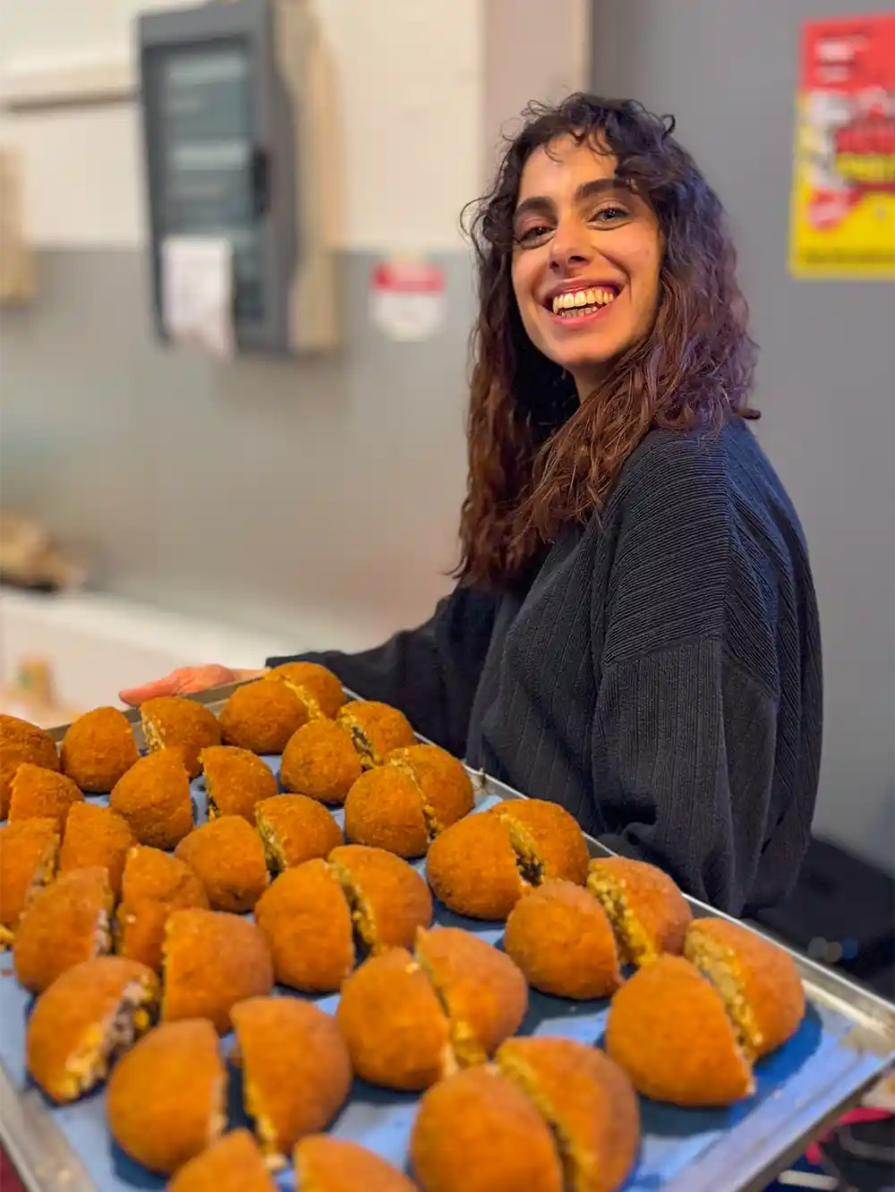 Gelato-Arancini-Gioia Eine lächelnde Frau hält ein Tablett mit runden, goldbraun gebratenen Snacks in der Hand, die aufgeschnitten sind, um die Füllung zu zeigen.