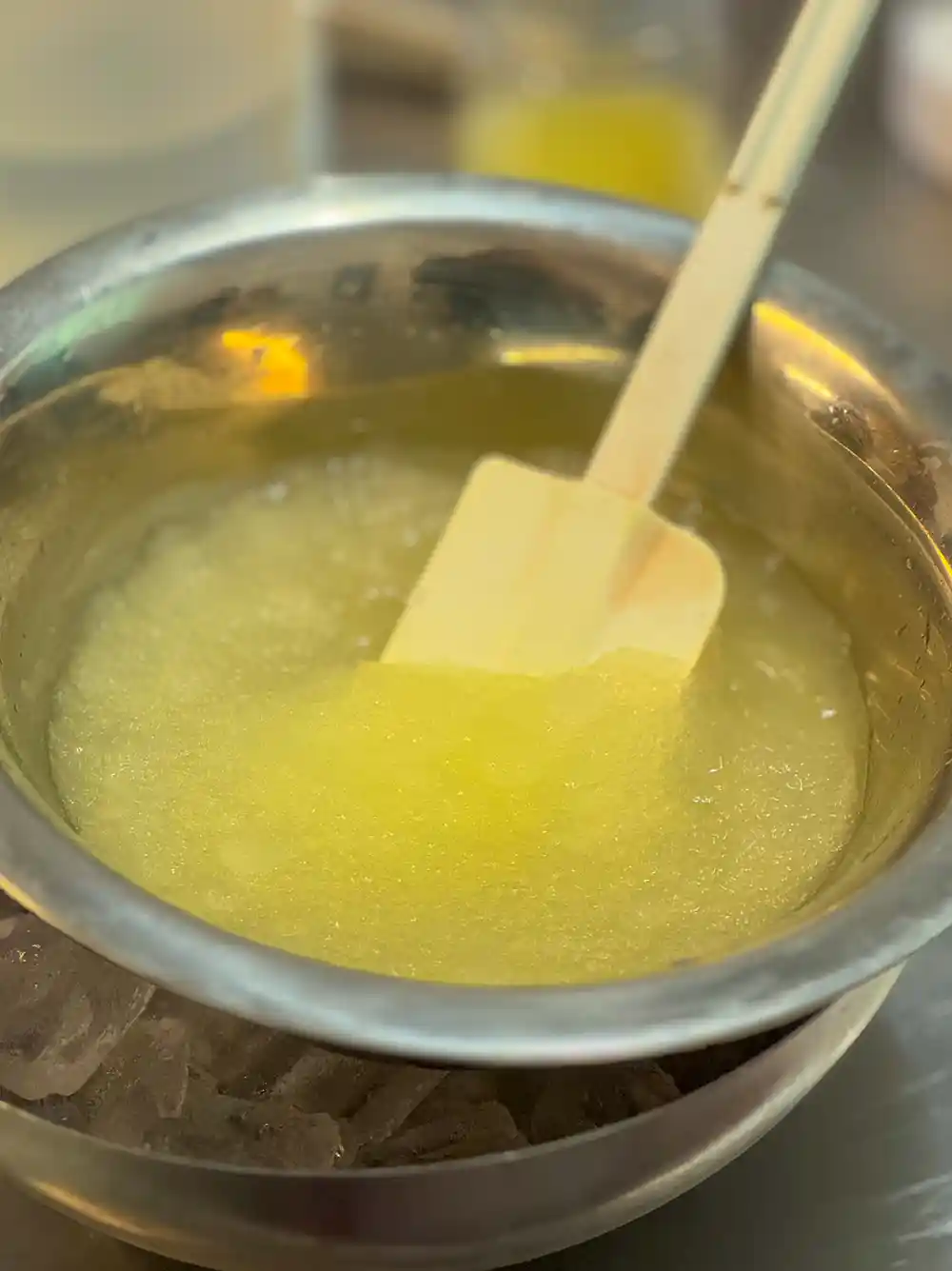 Italienisches-Granita-Herstellung-Duo- Ein Spatel rührt eine gelbe, matschige Masse in einer Metallschüssel, die über einer weiteren Schüssel mit Eis steht.