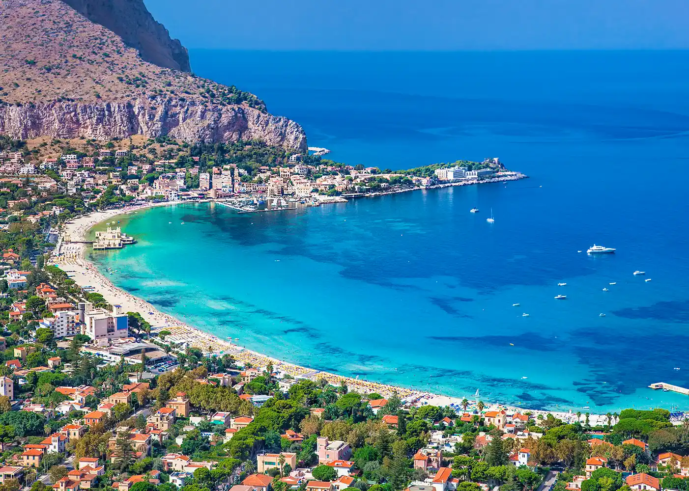 Luftaufnahme einer Küstenstadt mit türkisfarbenem Wasser, einem Sandstrand und grünen Hügeln unter einem klaren blauen Himmel.