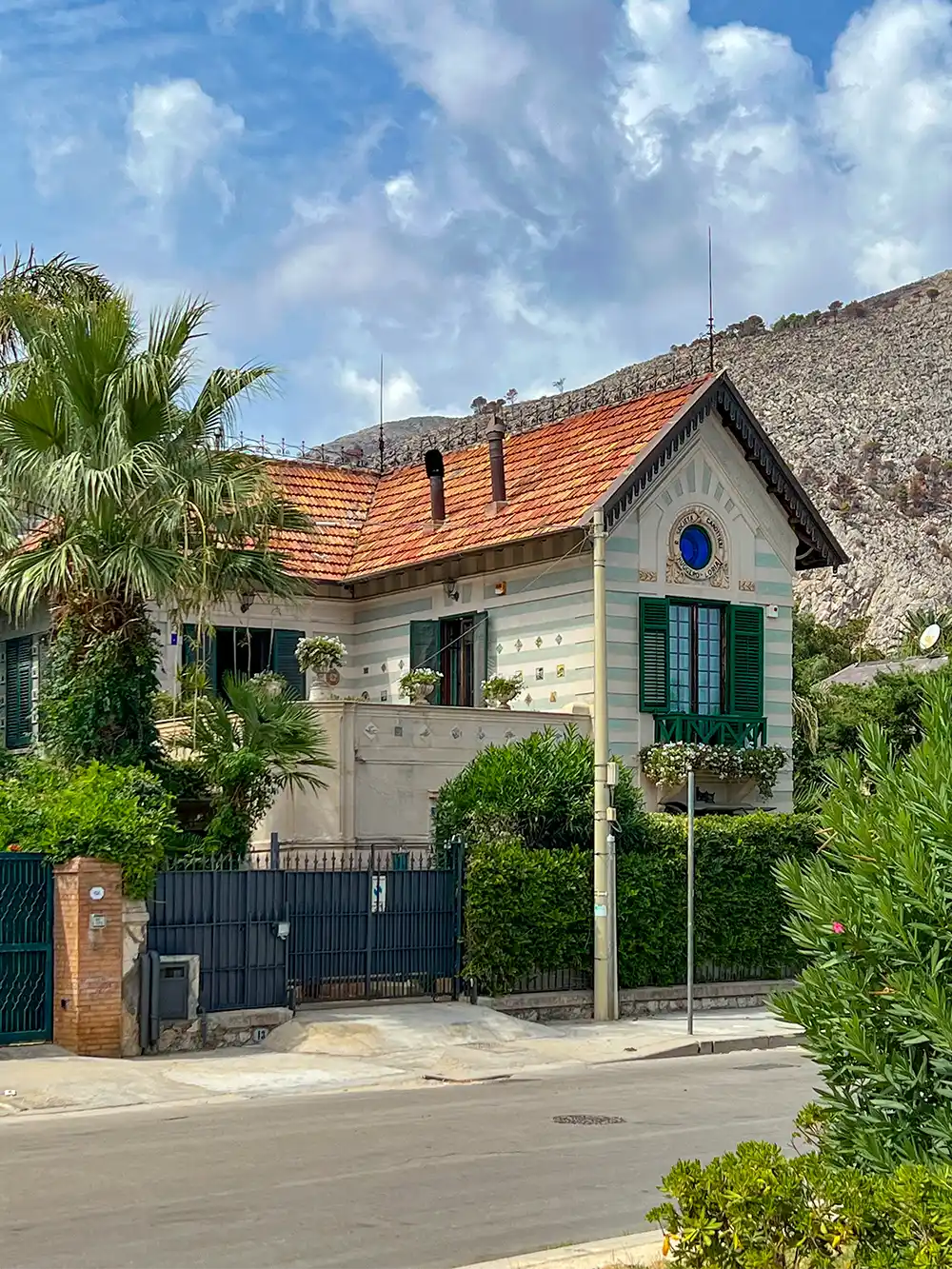 Ein zweistöckiges Haus mit rotem Dach, üppigem Grün und einem Berg im Hintergrund unter einem teilweise bewölkten Himmel.