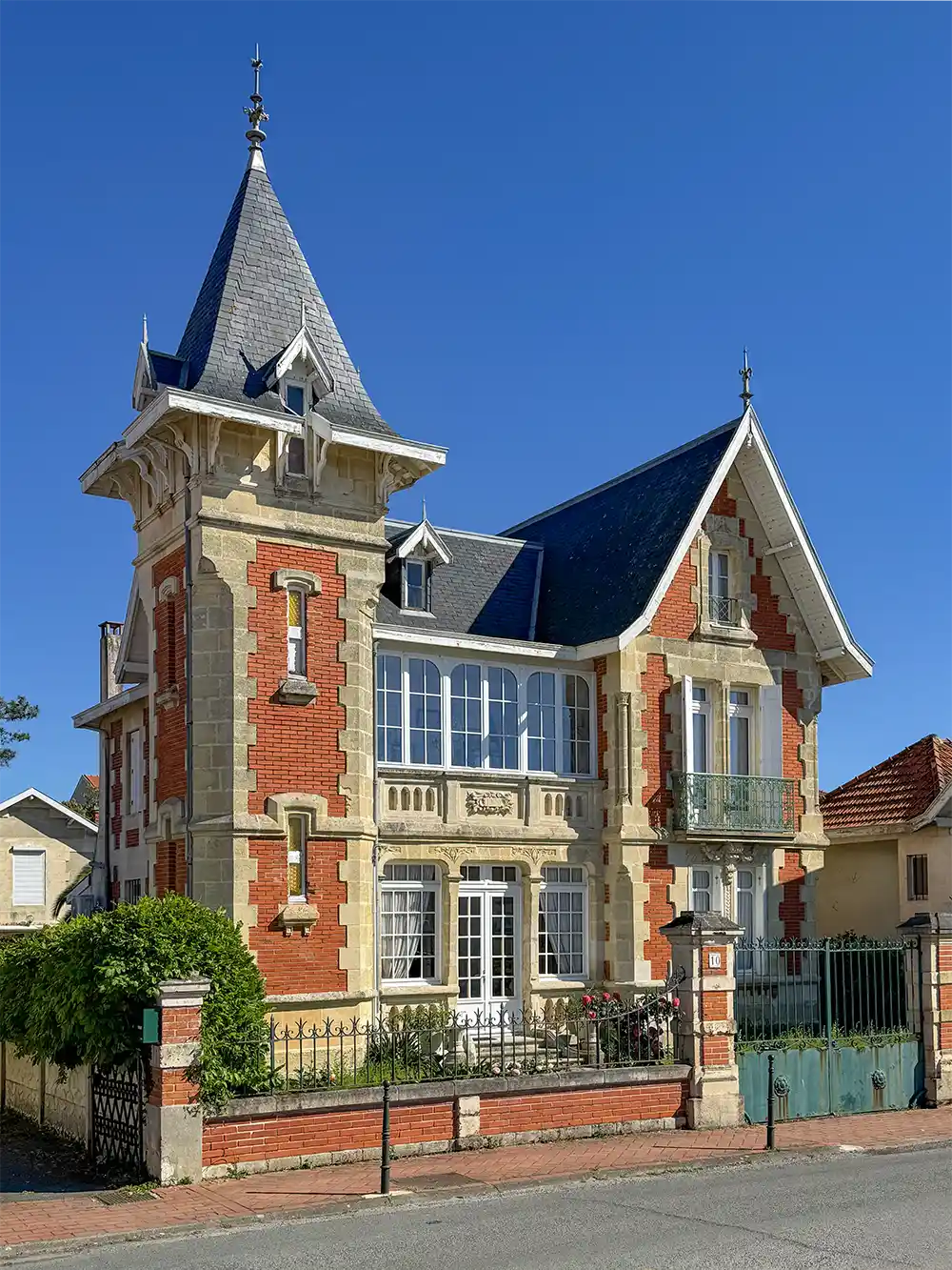 Soulac-Villa Ein reizvolles Haus aus Ziegeln und Stein mit einem hohen Türmchen, kunstvollen Verzierungen und einem grünen Tor unter einem klaren blauen Himmel.