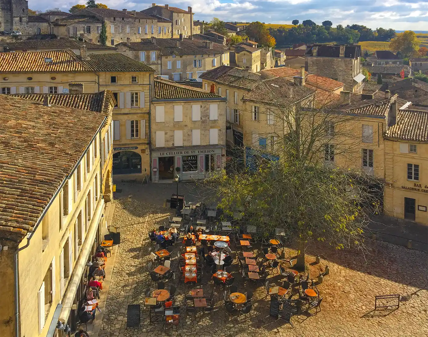 St-Emilion Ein Café mit Sitzplätzen im Freien auf einem kopfsteingepflasterten Platz, umgeben von sandfarbenen historischen Steingebäuden in einer sonnigen französischen Kleinstadt.