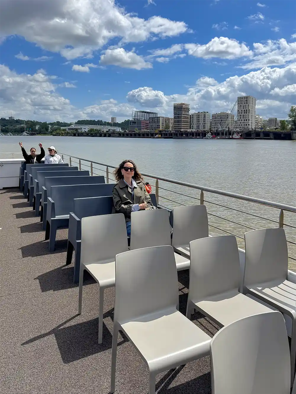 Bordeaux-Geheimtipp-Bootsfahrt-Garonne Eine Frau sitzt allein auf einer Fähre, umgeben von leeren Stühlen; im Hintergrund winken zwei Menschen am Flussufer.