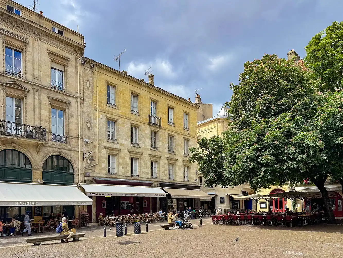 Bordeaux-Sehenswuerdigkeit-Place-Saint-Pierre Ein sonnendurchfluteter europäischer Platz mit Straßencafés, sitzenden Menschen und großen Bäumen neben historischen Gebäuden.