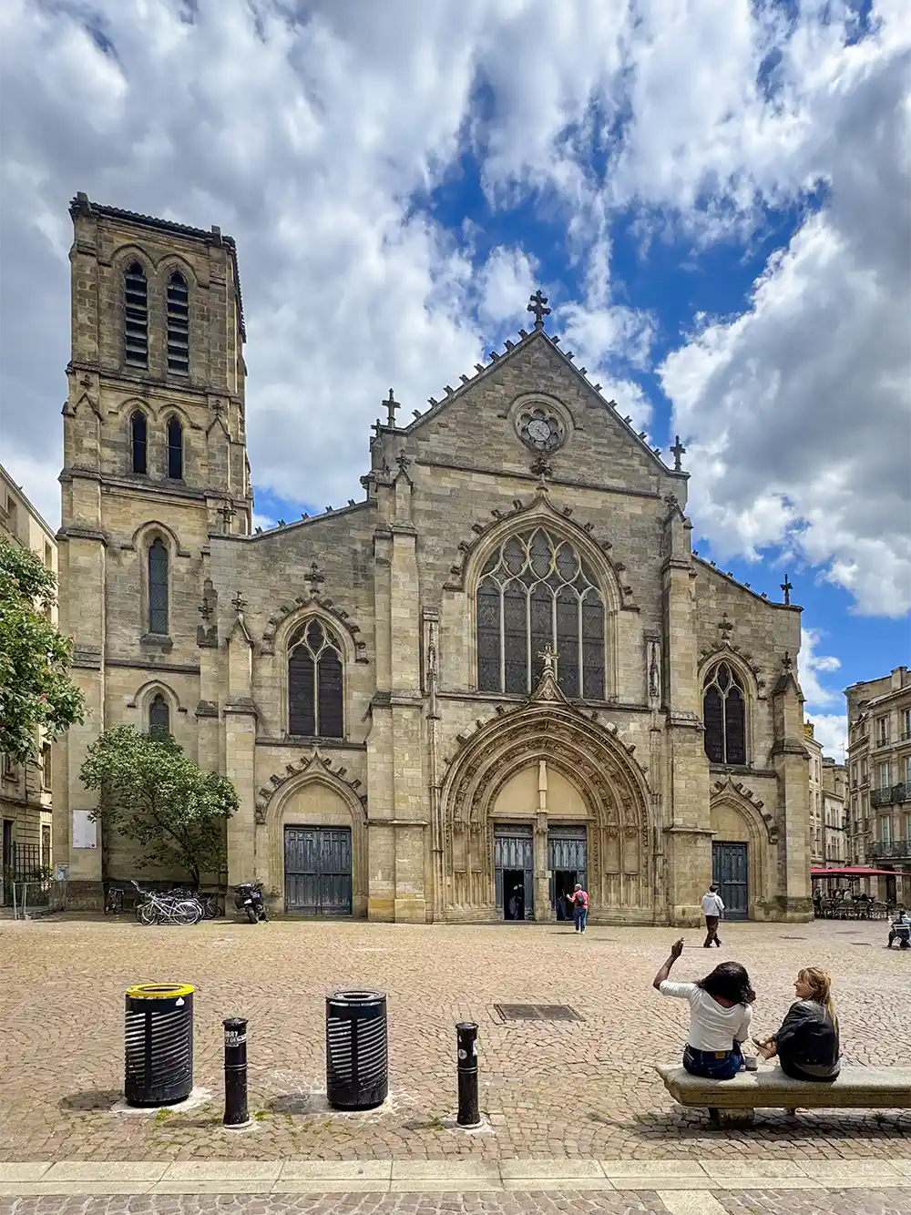 Bordeaux-Sehenswuerdigkeit-Saint-Pierre Eine historische Steinkirche mit einem hohen Glockenturm, Menschen, die auf einer Bank sitzen, und einem bewölkten Himmel darüber.