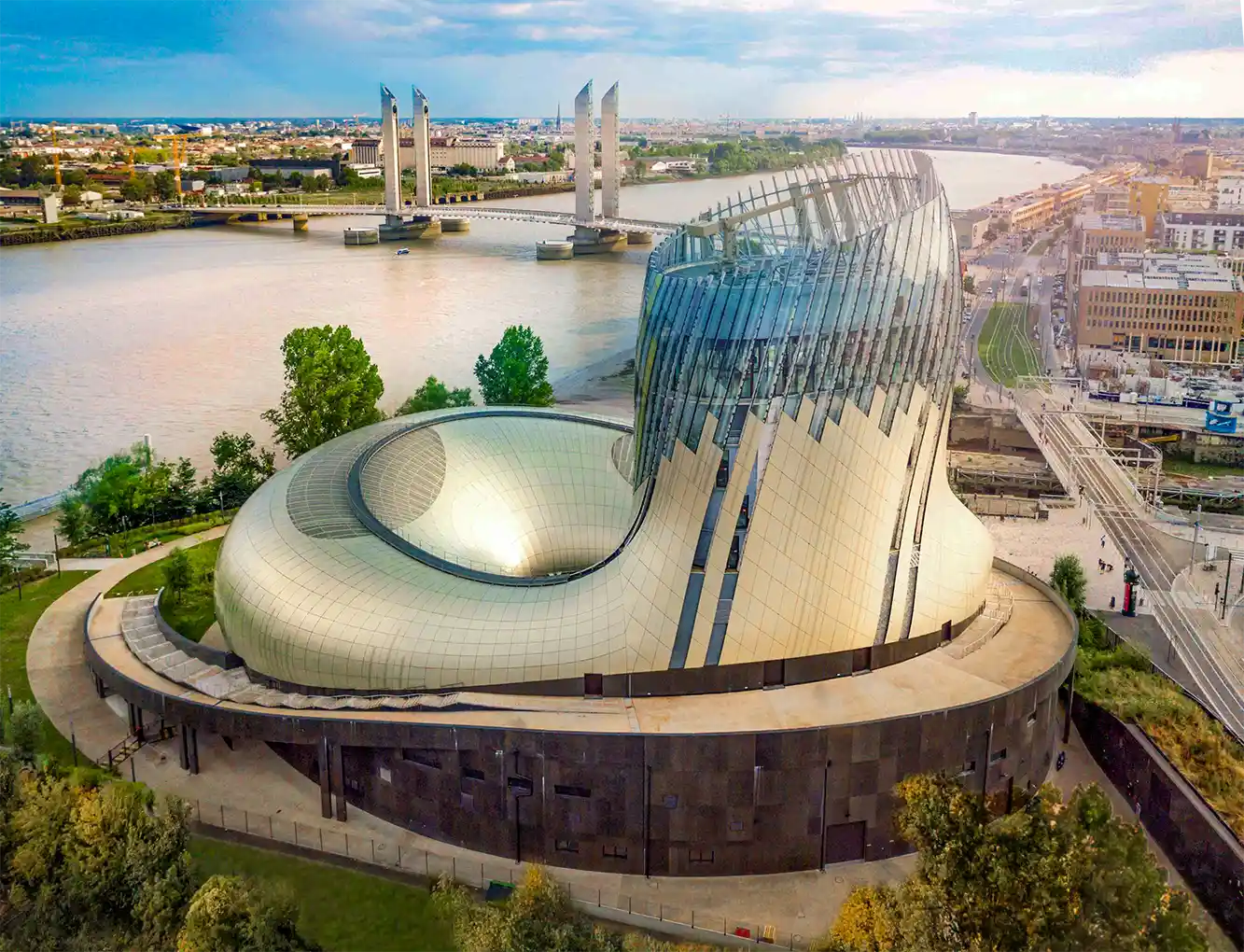 Cite-du-vin-Bordeaux Ein futuristisches, geschwungenes Gebäude aus Glas und Metall am Flussufer, im Hintergrund eine Hubbrücke.