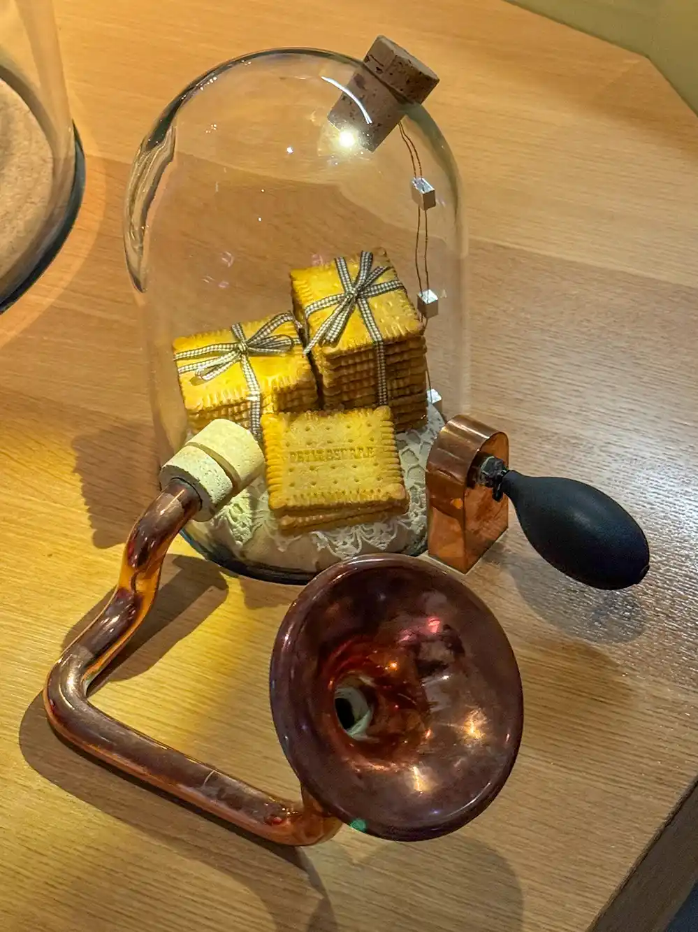 Vier übereinander gestapelte Kekse unter einer Glasglocke, neben einem alten Kupferhorn und einem schwarzen Gummiball auf einer Holzunterlage.