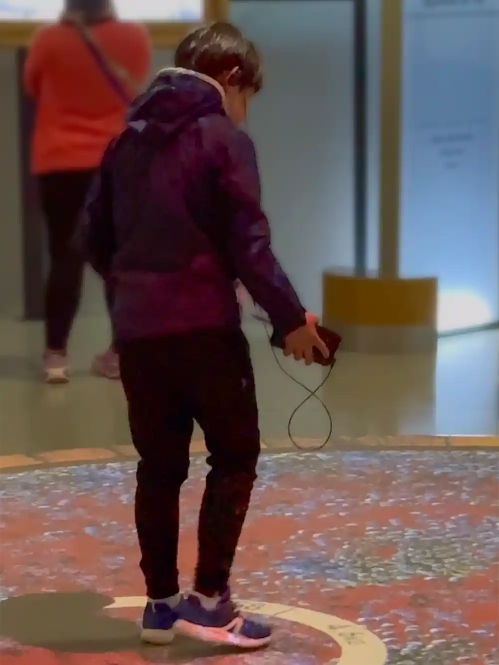 Ein Junge steht auf einem gemusterten Boden und hält ein Gerät mit einem Kabel in der Hand, während im Hintergrund eine Frau zu sehen ist.