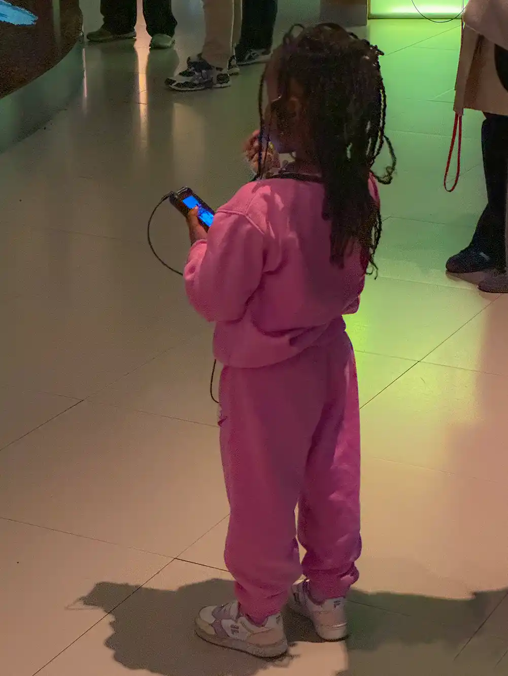 Ein kleines Kind in einem rosa Outfit steht drinnen, hält ein Gerät in der Hand und trägt Kopfhörer.