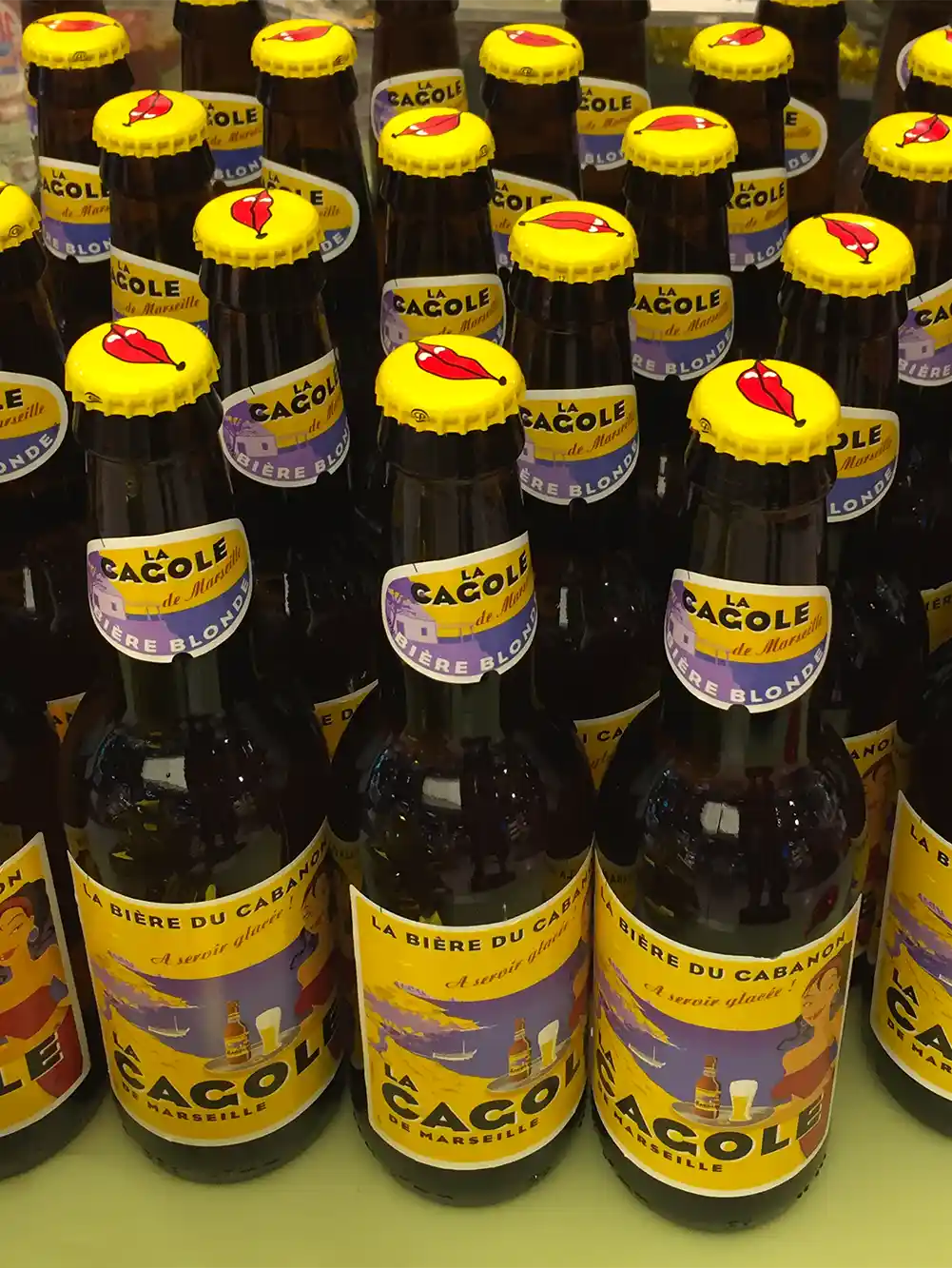 Mehrere Flaschen La Cagole-Bier mit gelben Deckeln und roten Lippen stehen dicht beieinander.
