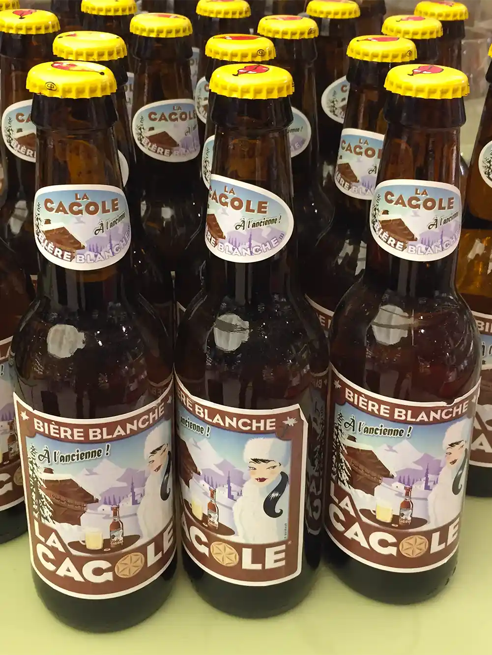 Mehrere Flaschen La Cacole Bière Blanche mit gelben Verschlüssen stehen zusammen in einem Regal.