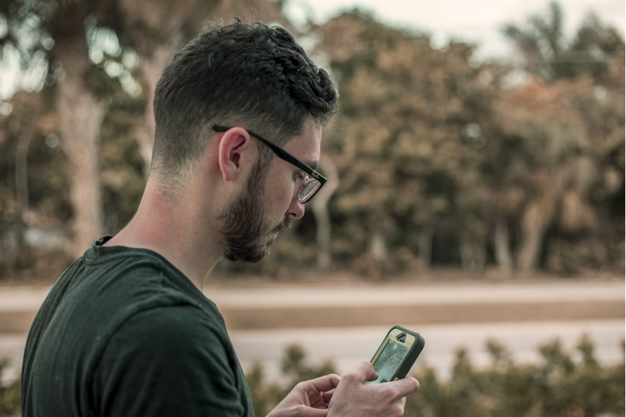 e-sim-telefonieren-2 Ein Mann mit Brille steht im Freien und schaut auf sein Smartphone, im Hintergrund sind Bäume und eine Straße zu sehen.