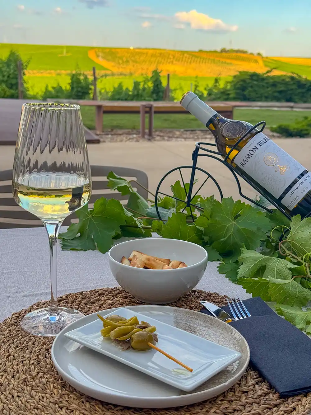 Bodega-Ramon-Bilbao Ein Glas Weißwein, Oliven und Brot auf einem Tisch im Freien, im Hintergrund der Blick auf die Weinberge.