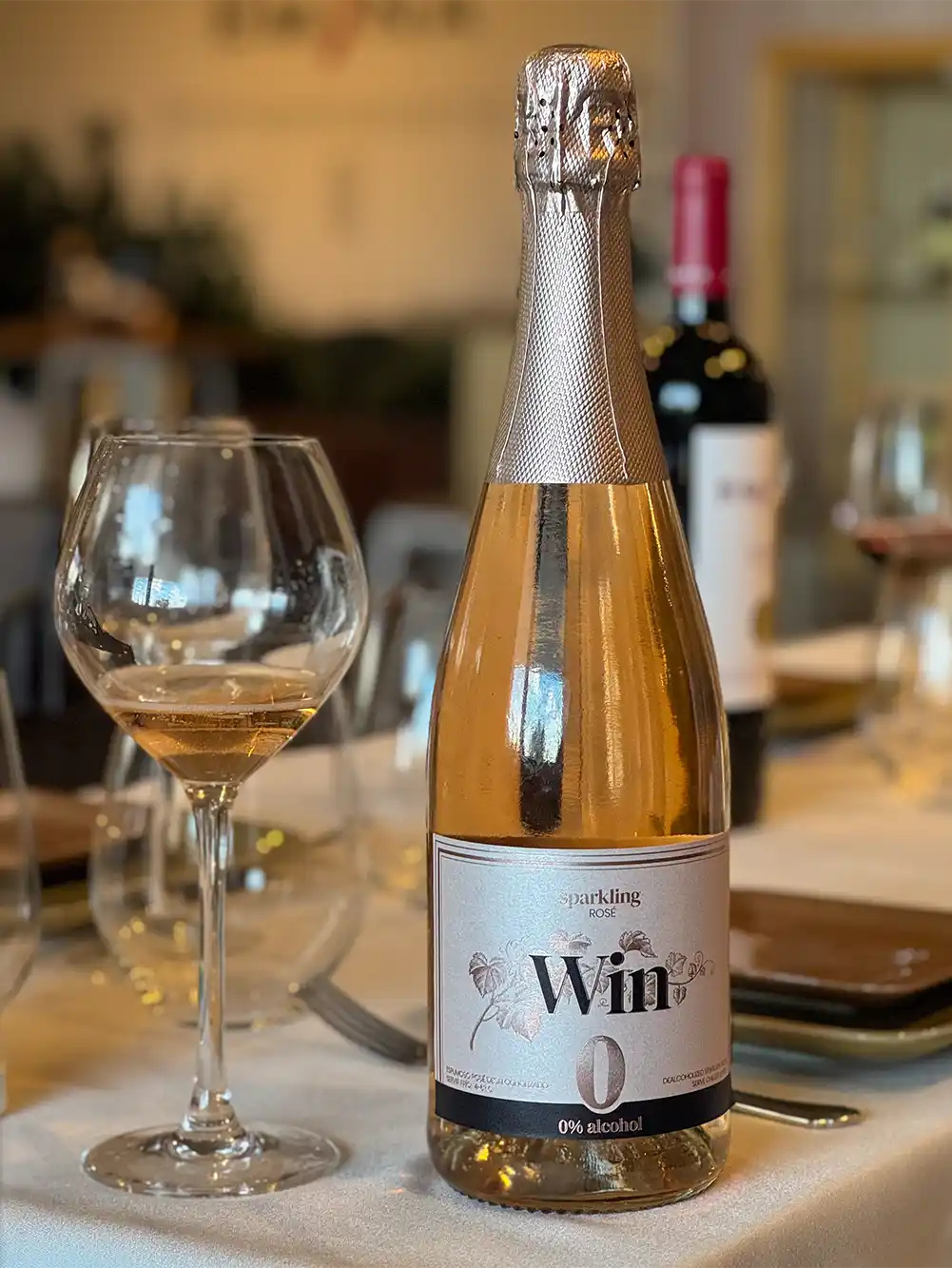 Bodega-Win-Emina-alkoholfreier-Sekt Eine Flasche Rosé-Sekt und ein gefülltes Glas auf einem gedeckten Esstisch, mit der Aufschrift „0 % Alkohol“.