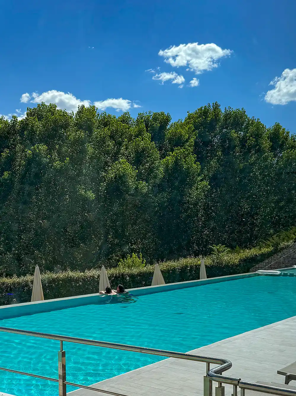 Castilla-Termal-Pool Ein klarer Pool, in dem zwei Personen schwimmen, im Hintergrund grüne Bäume, darüber ein blauer Himmel mit ein paar Wolken.