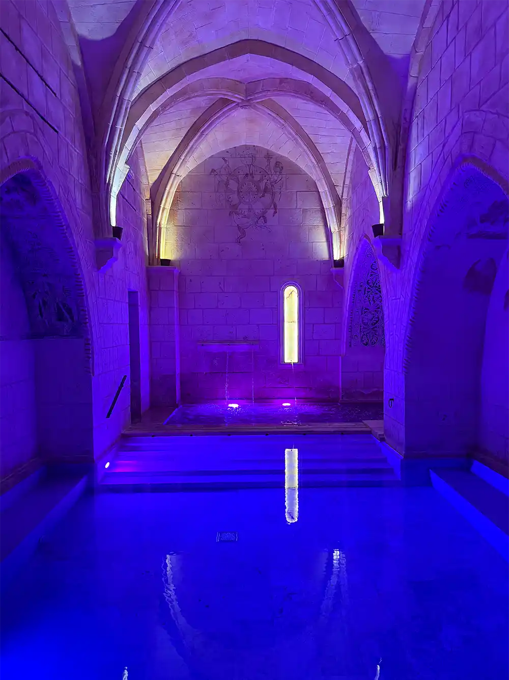 Castilla-Termal-Spa Bad unter einer gewölbten gotischen Steindecke, beleuchtet mit violettem und blauem Licht, das sich auf dem Wasser spiegelt.