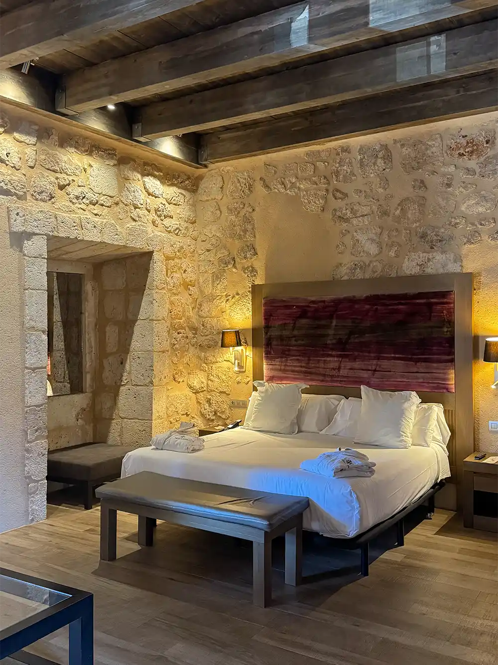Castilla-Termal-Zimmer Gemütliches Hotelzimmer mit Steinwänden, einem Bett, Holzbalken, Bänken und warmer Beleuchtung.
